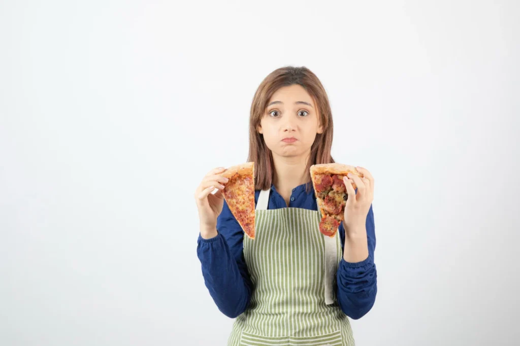 Young woman in apron holding two slices of pizza with concerned expression, illustrating dilemma about eating processed foods that damages skin health