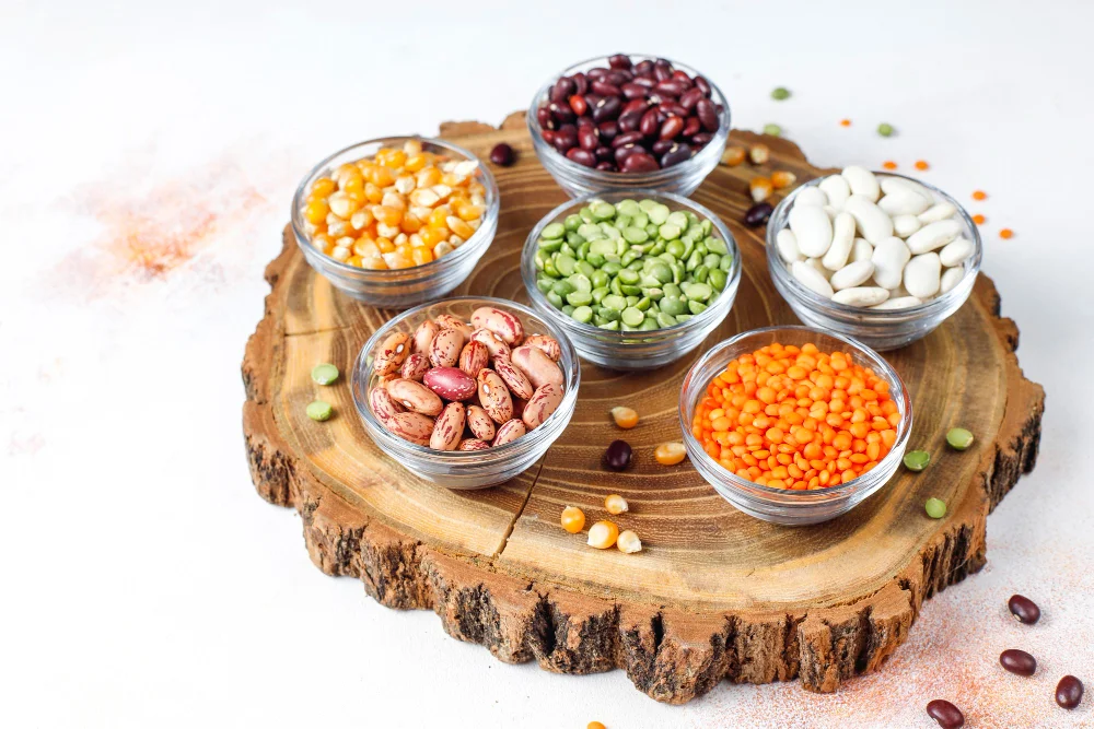 Six glass bowls arranged on a wooden cutting board displaying various colorful beans and legumes including yellow split peas, red kidney beans, green split peas, white beans, pinto beans, and orange lentils