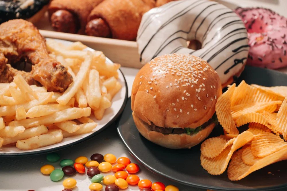 Assortment of processed and fast foods including glazed donuts, fried chicken, french fries, a sesame seed burger, potato chips, and colorful candy displayed on plates