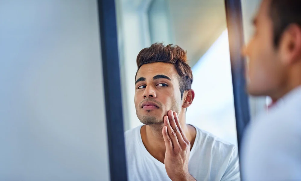 Young man examining his facial skin in mirror checking for side effects of daily masturbation